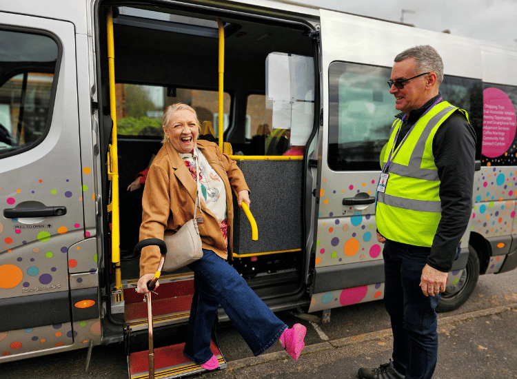 An older woman happily disembarking an accessible minibus, assisted by a uniformed Communities 1st staff member, showing transport services.