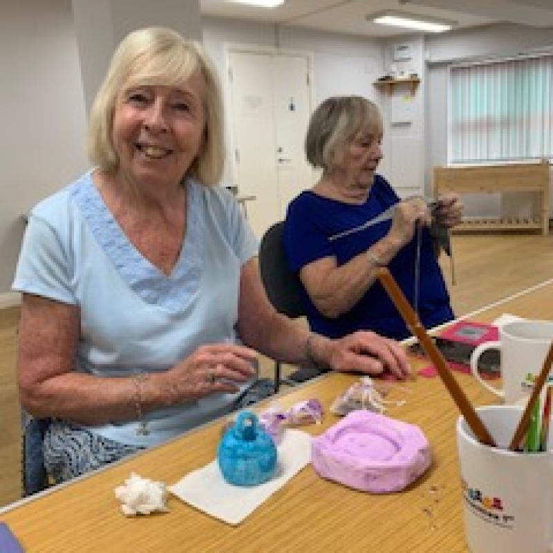 Smiling lady painting a small clay sculpture. The lady sitting next to her is knitting